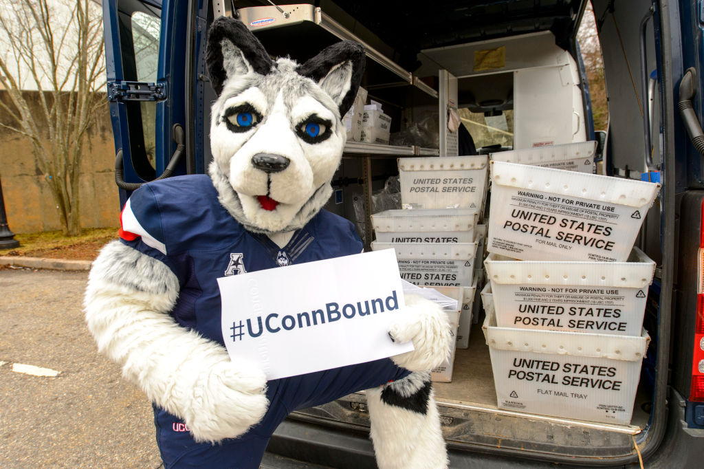 Jonathan the Husky stands next to a UConn Mail Services truck and shows off admissions acceptance packets.